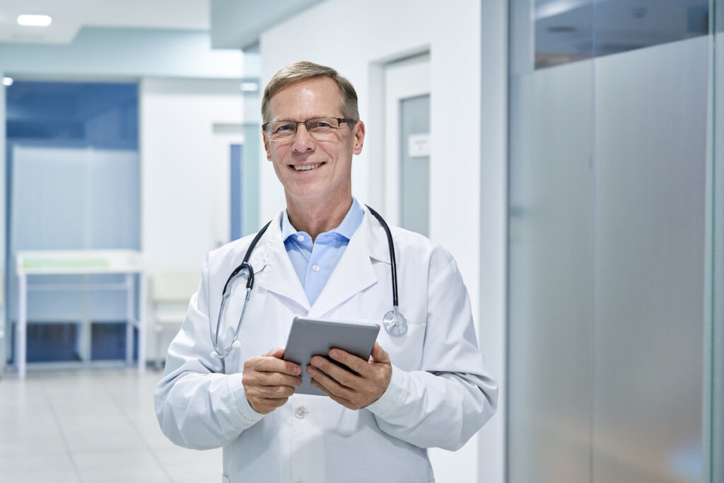 Smiling middle-aged doctor holding a digital tablet standing in hospital looking at camera.