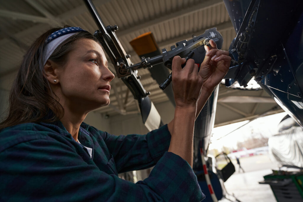 Brunette, Caucasian, middle-aged woman using a flashlight while analyzing airplane parts in hangar in aviation garage.