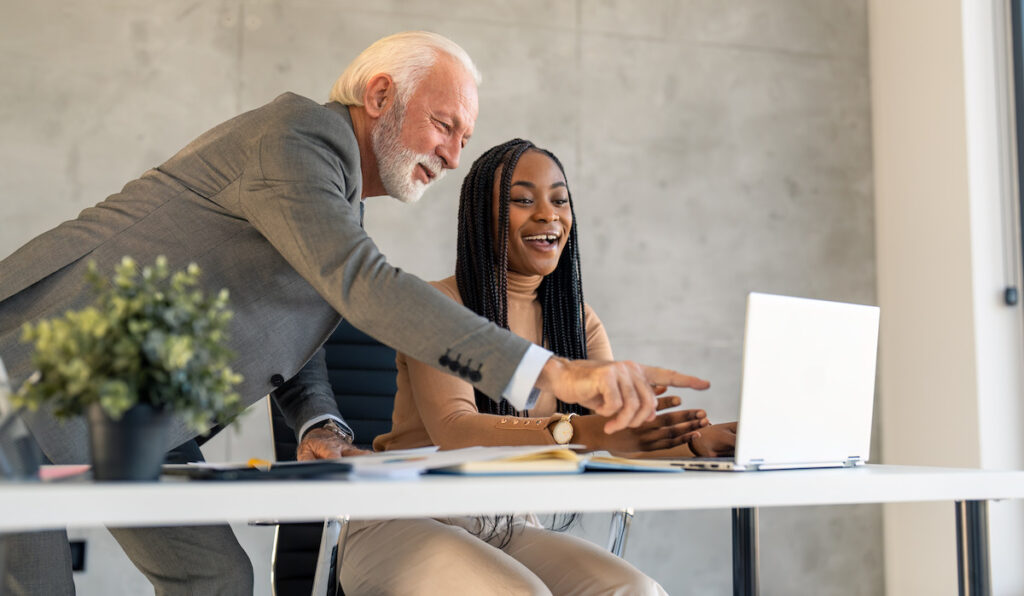 Senior male businessman executive manager mentor explaining working operations to female African American colleague coworker intern using laptop in modern office room.